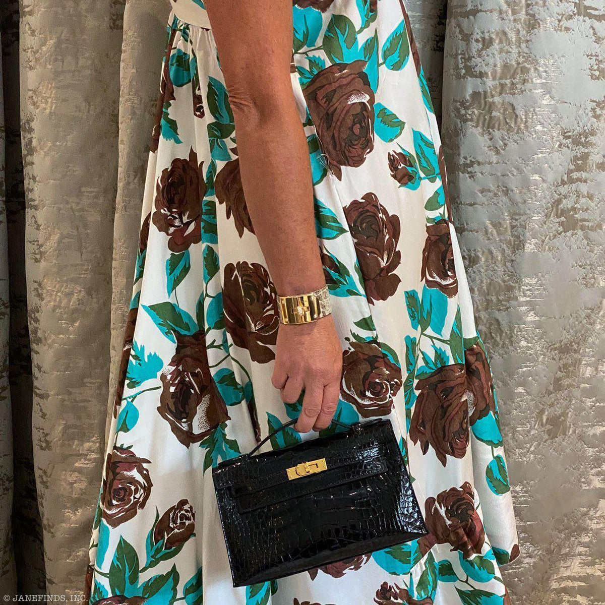 Person wearing a floral dress with brown roses holding a black handbag against a textured wall.